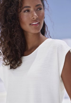 White textured top with short sleeves, featuring a rounded neckline. Model has curly hair, posing against a light background.