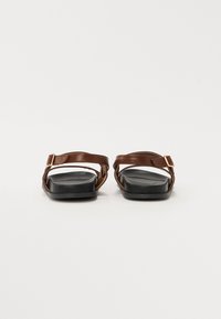 Sandalias de cuero marrón con un acento de hebilla, suela plana negra y diseño de tiras cruzadas. Textura suave con una apariencia minimalista.