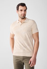 Beige poloing, textúrált pálmalevél mintával, rövid ujjú és galléros nyakkal. Olíva zöld nadrággal párosítva.