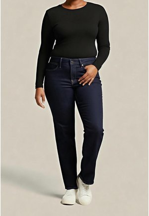 Woman wearing a black long-sleeve top, dark blue jeans, and white shoes standing against a plain light background.