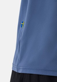 Blaues Sportshirt aus einem strukturierten, atmungsaktiven Material. Verfügt über ein kleines schwedisches Flaggen-Patch am Saum und einen schwarzen unteren Rand.