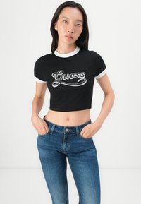 Svart kortermet t-skjorte med hvit ribbet krage og mansjetter; har "Guess" logo i stilisert hvit skrift; kombinert med blå høyt midje jeans.