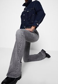 Chaqueta vaquera en azul oscuro con botones plateados y dos bolsillos delanteros, combinada con jeans acampanados en gris claro y botas negras gruesas.