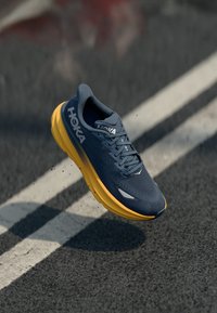 HOKA løpesko har en marineblå mesh-overdel med et teksturert mønster og en lys gul polstret såle. Lett design med reflekterende detaljer.