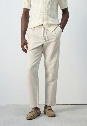 Homme portant un pantalon écru avec cordon de serrage, une chemise beige tissée à manches courtes et des mocassins couleur fauve, debout les mains dans les poches.