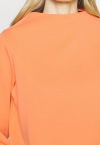 Oranssi huppari, jossa on pyöreä pääntie, pitkät hihat ja sileä, hieman teksturoitu kangas. Minimalistinen muotoilu ilman näkyviä koristeita.