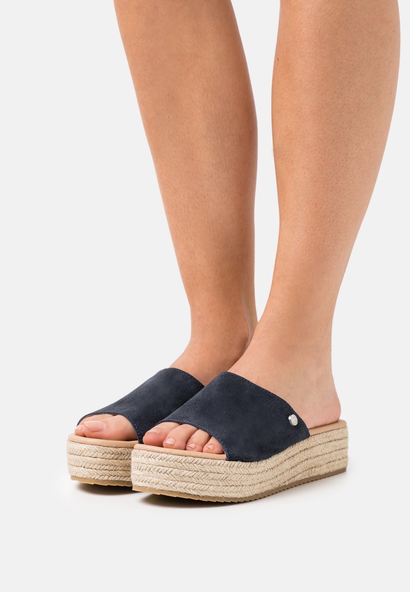 Sandales mules en daim bleu marine à bouts ouverts, avec une semelle en jute tissée et un léger accent métallique sur le côté. Design à plateforme surélevée.