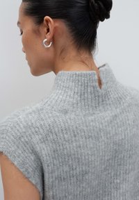 Frau mit dunklem Haar, hochgesteckt, trägt einen ärmellosen, hochgeschlossen, flauschigen grauen Strickpullover und silberne Creolen, steht mit dem Rücken zu einem schlichten Hintergrund.