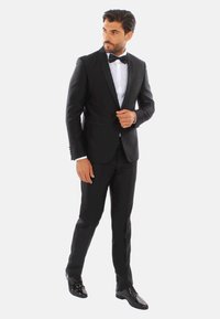 Tuxedo nero con accenti in satin, dotato di giacca aderente, pantaloni abbinati e camicia bianca. Scarpe eleganti nere lucide.