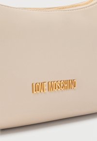 Επιφάνεια από μπεζ δέρμα με ανάγλυφο χρυσό λογότυπο "LOVE MOSCHINO" και διακριτική λεπτομέρεια ραφής στην επάνω άκρη.