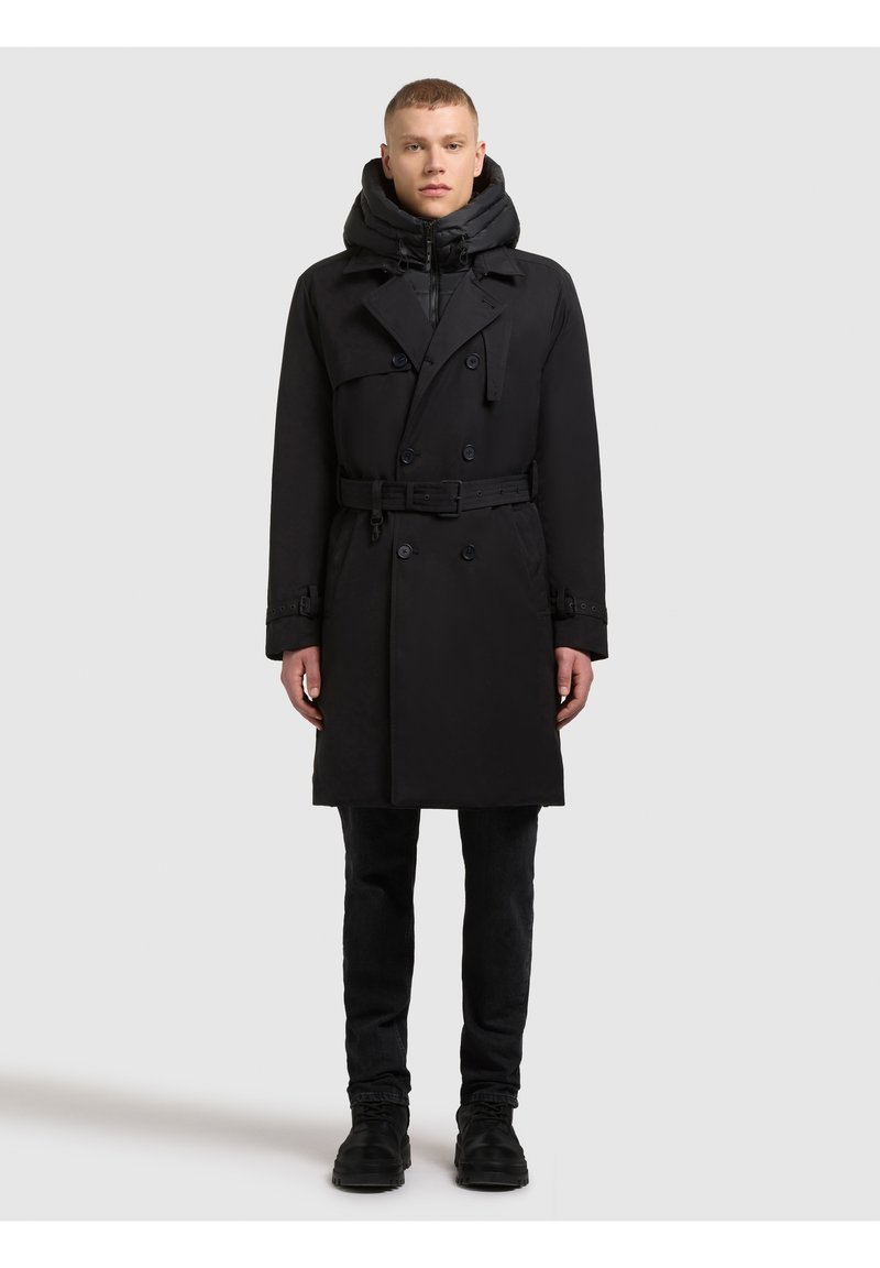 Czarny płaszcz typu trench z podwójnym zapięciem, regulowanym paskiem i ocieplaną kapturem. Noszony na czarnych dopasowanych spodniach i butach.