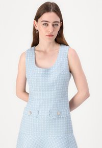 Robe en tweed bleu clair avec un décolleté carré, sans manches, et poches avant décoratives avec des éléments ornés. Tissu texturé.