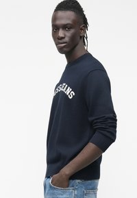 Tumši zilss džemperis ar apaļu apkakli, priekšpusē balti burti "JEANS", ar riboto aproču un jostasvietas apdari, kopā ar gaiši zilām džinsām.