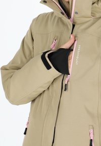 Giacca impermeabile beige con zip rosa, multiple tasche e polsini regolabili con velcro nero. Tessuto liscio e durevole.