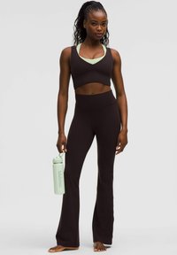 Zwarte sportieve crop top en hoog-wijde flared leggings; model houdt een lichtgroene waterfles vast; zachte textuur en slimfit ontwerp.