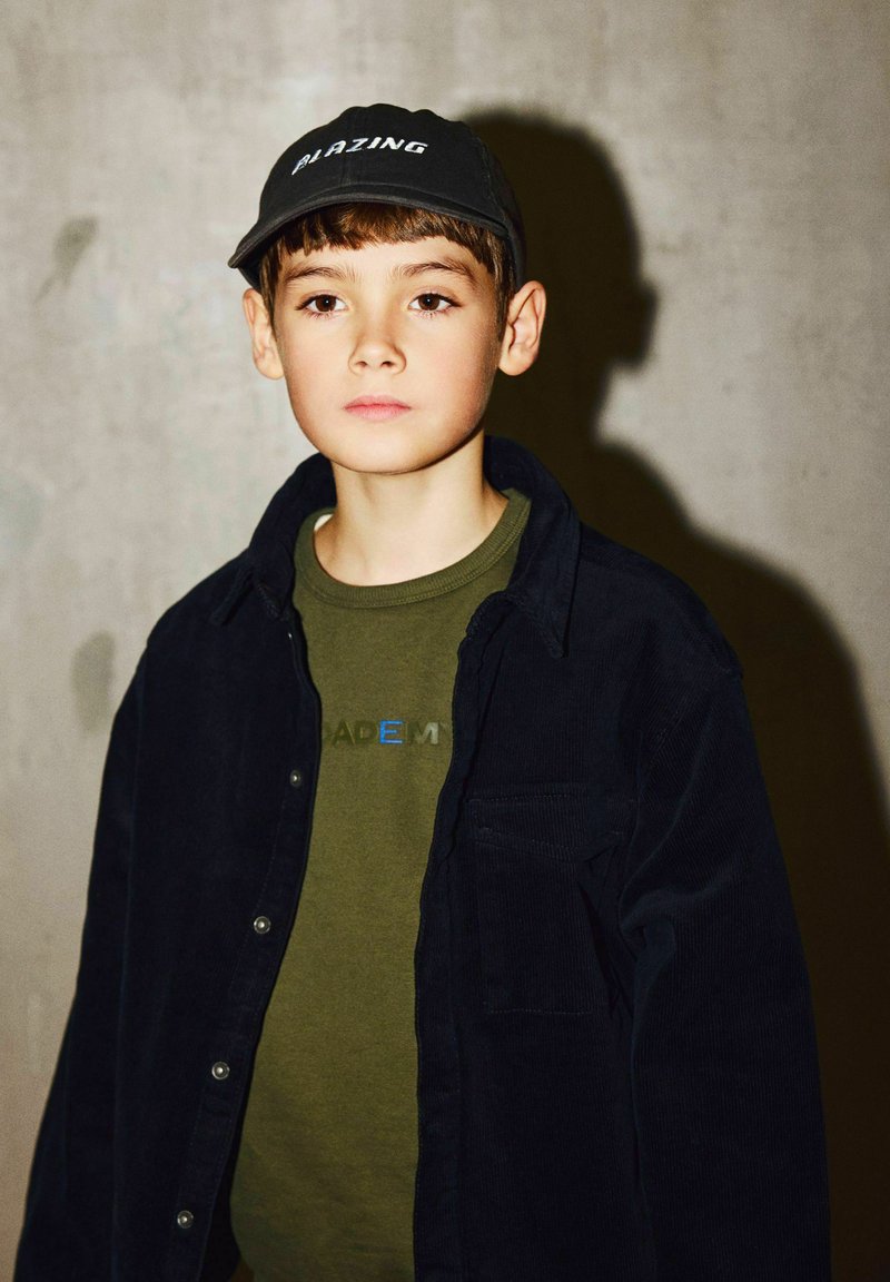 Garçon portant une casquette noire, un t-shirt vert "BLAZING," et une veste sombre se tient contre un mur beige en projetant une ombre.