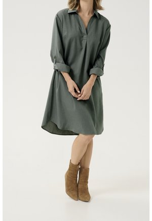 Groene overhemdjurk tot op de knie met een kraag, opgerolde mouwen en een uitlopende onderkant. Draagt bruine enkellaarzen. Gestructureerde stof.