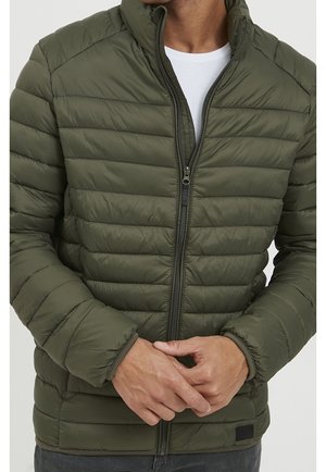 Mężczyzna w oliwkowozielonej pikowanej kurtce puchowej zapiętej do połowy na białą koszulę, ręce splecione z przodu, widoczne szare spodnie.