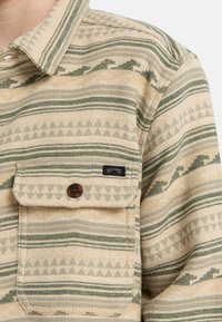 Camicia button-up tan con motivi geometrici verdi e crema. Presenta una tasca sul petto e un'etichetta con logo. Tessuto lavorato testurizzato.