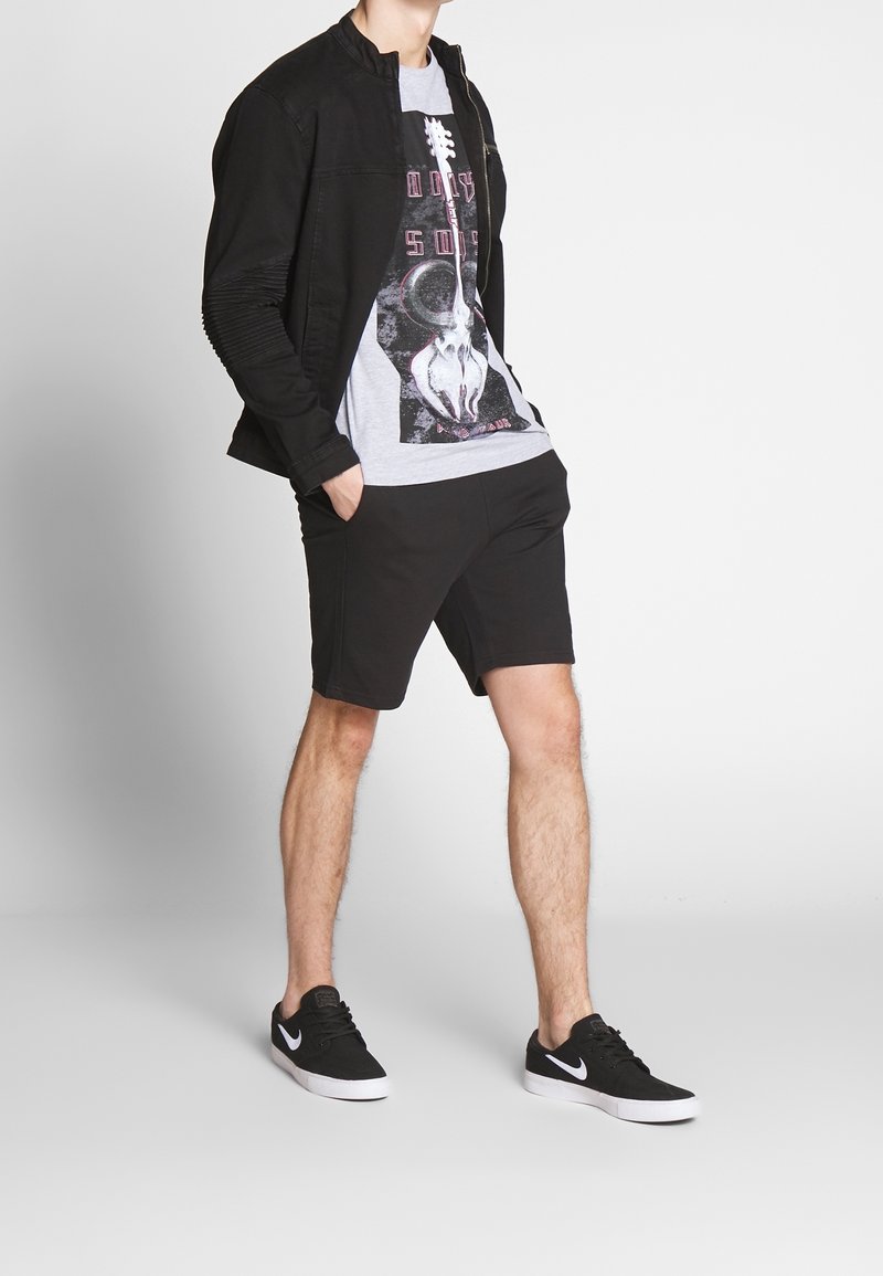 Veste en jean noire avec détails côtelés aux manches, t-shirt gris à motif crâne, short noir, et baskets Nike noires avec des accents blancs.