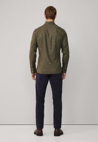 Man in een olijfgroene blouse met lange mouwen en kleine, roodachtige patronen, marineblauwe broek en bruine schoenen, staande met zijn gezicht naar een eenvoudige grijze muur.