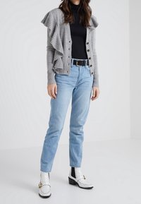 Cárdigan gris acanalado con detalles de volantes, camiseta negra, vaqueros azul claro y botines blancos con herrajes dorados y tacones negros.