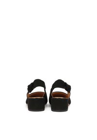 Black suede block-heeled sandals featuring a knotted strap detail and an open back. The insoles are tan, providing contrast to the outer material.