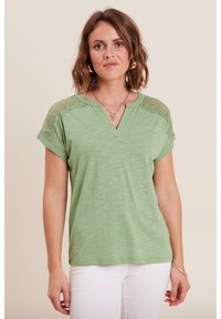 Grünes, kurzärmliges Oberteil mit V-Ausschnitt, strukturiertem Stoff und Spitzenakzenten an den Schultern. Kombiniert mit weißen Hosen.