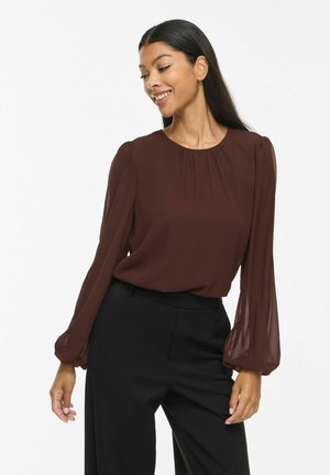 Femme aux longs cheveux foncés, portant un chemisier marron à manches longues et un pantalon noir, souriante et regardant vers le bas sur le côté.