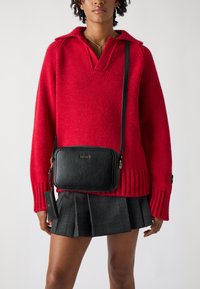 Schwarze Umhängetasche mit glatter Textur, rechteckiger Form, goldenem Logo und verstellbarem Riemen, kombiniert mit einem roten Strickpullover und einem karierten Rock.