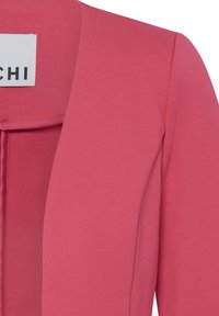 Gros plan sur la manche et le revers d'un blazer rose avec une étiquette intérieure blanche partiellement visible portant le texte "CHI".