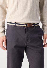 Marineblaue Baumwollhose mit einem hellbeigen Pullover. Ein gestreifter Gürtel mit marineblauen, weißen und braunen Akzenten, befestigt mit einer goldfarbenen Schnalle.