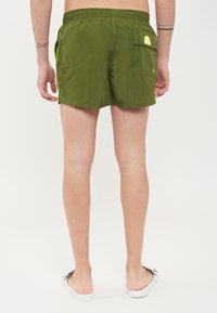 Shorts de bain verts avec une poche arrière, une taille élastique et des bandes latérales jaunes contrastantes. Le matériau semble léger et lisse.