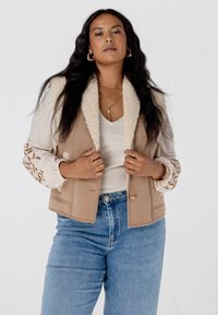 Beige gilet met schapenvachtkraag en een zachte textuur, versierd met bloemembroidery op lange mouwen, gecombineerd met lichtblauwe jeans.