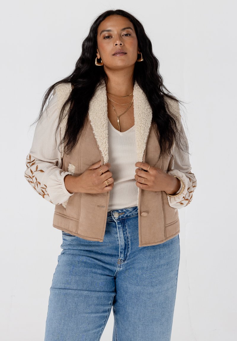 Beige gilet met schapenvachtkraag en een zachte textuur, versierd met bloemembroidery op lange mouwen, gecombineerd met lichtblauwe jeans.