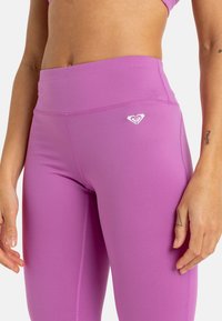 Vaaleanpunaiset korkeavyötäröiset legginsit, jotka on valmistettu joustavasta kankaasta ja joissa on sileä pinta sekä pieni valkoinen logo. Näkyvät saumat jaloissa.