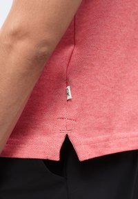 Gros plan d'une personne portant une chemise texturée rose avec une petite étiquette blanche à logo près de la couture latérale et un pantalon noir.