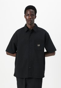 Camisa negra de manga corta y botones con una tela texturizada, un bolsillo en el pecho que presenta un logo dorado de "GAP" y un diseño de ajuste relajado.