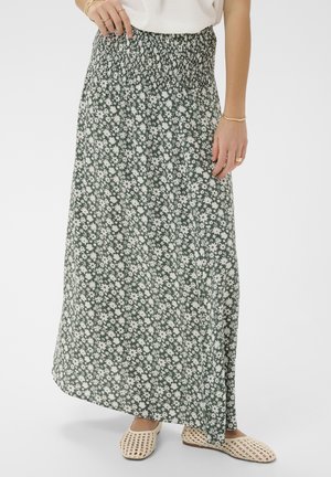 Frau trägt einen langen grünen Rock mit Blumenmuster und elastischem Bund, weißes Oberteil, goldenen Schmuck und cremefarbene flache gewebte Schuhe, steht vor weißem Hintergrund.