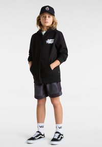 Sudadera con cremallera negra con el logotipo de Vans, combinada con pantalones cortos grises estampados, zapatillas negras de Vans y calcetines blancos con la marca Vans.