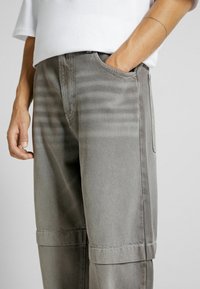 Man med löst sittande grå byxor och en vit skjorta, en hand i fickan, en beskuren armtatuering synlig på underarmen.