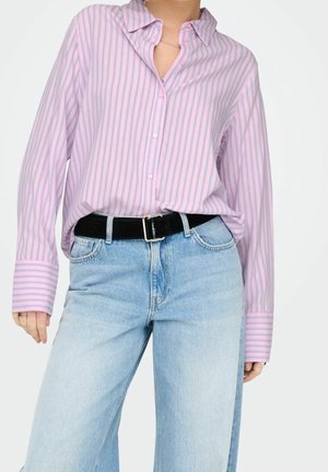 Personne portant une chemise ample rose à rayures boutonnée rentrée dans un jean taille haute bleu clair avec une ceinture noire.