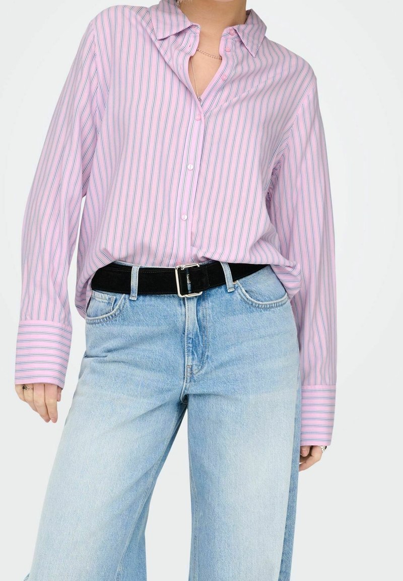 Personne portant une chemise ample rose à rayures boutonnée rentrée dans un jean taille haute bleu clair avec une ceinture noire.