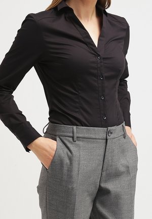 Femme portant une chemise noire moulante boutonnée rentrée dans un pantalon gris taille haute et ajusté, les mains dans les poches.