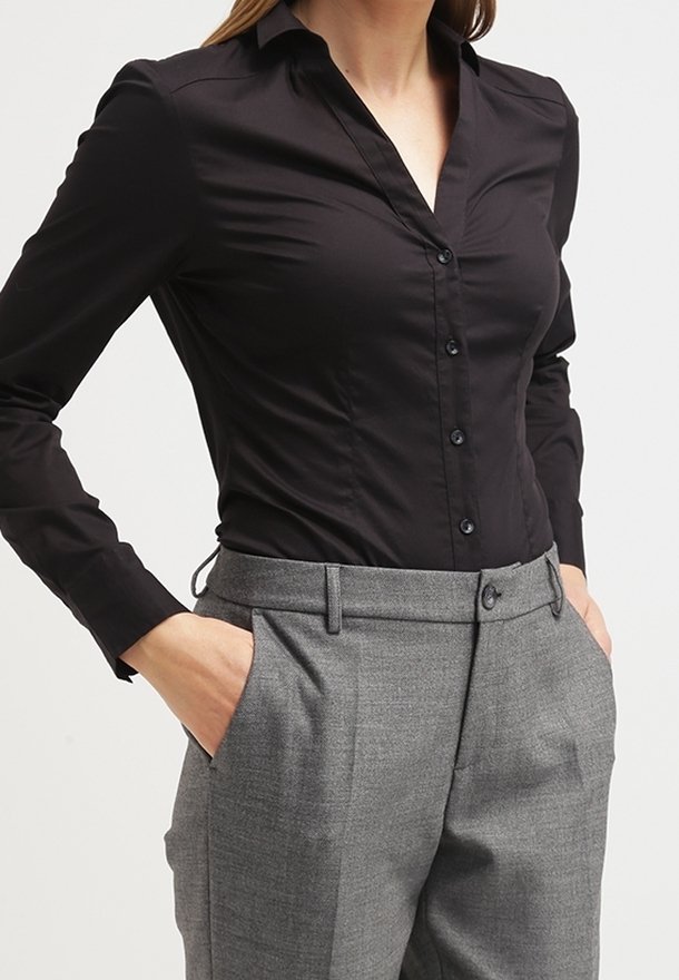 Femme portant une chemise noire moulante boutonnée rentrée dans un pantalon gris taille haute et ajusté, les mains dans les poches.