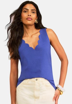 Žena nosí modrý tielkový top so zvlneným výstrihom, krémové nohavice, zlaté kruhové náušnice a masívne zlaté a biele náramky.