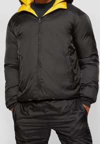 Person klädd i en svart puffjacka med gul insida och matchande svarta byxor, med händerna i fickorna, mot en enfärgad bakgrund.