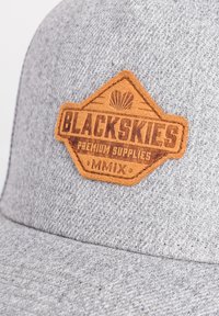 Grauerm Hut mit einem braunen Lederpatch, auf dem der Text "BLACKSKIES PREMIUM SUPPLIES MMIX" und ein Muschel-Design zu sehen sind. Strukturiertem Stoff.