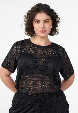 Femme aux cheveux bouclés portant un haut en dentelle noire à motifs et un short assorti, debout, les mains dans le dos, sur fond blanc.
