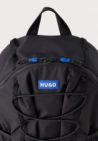 Juodas kuprinė su tekstūruota medžiaga, su mėlynomis užtrauktuko kilpomis ir mėlynu logotipo pleistru; priekinėje dalyje yra elastingos virvelės detalė.